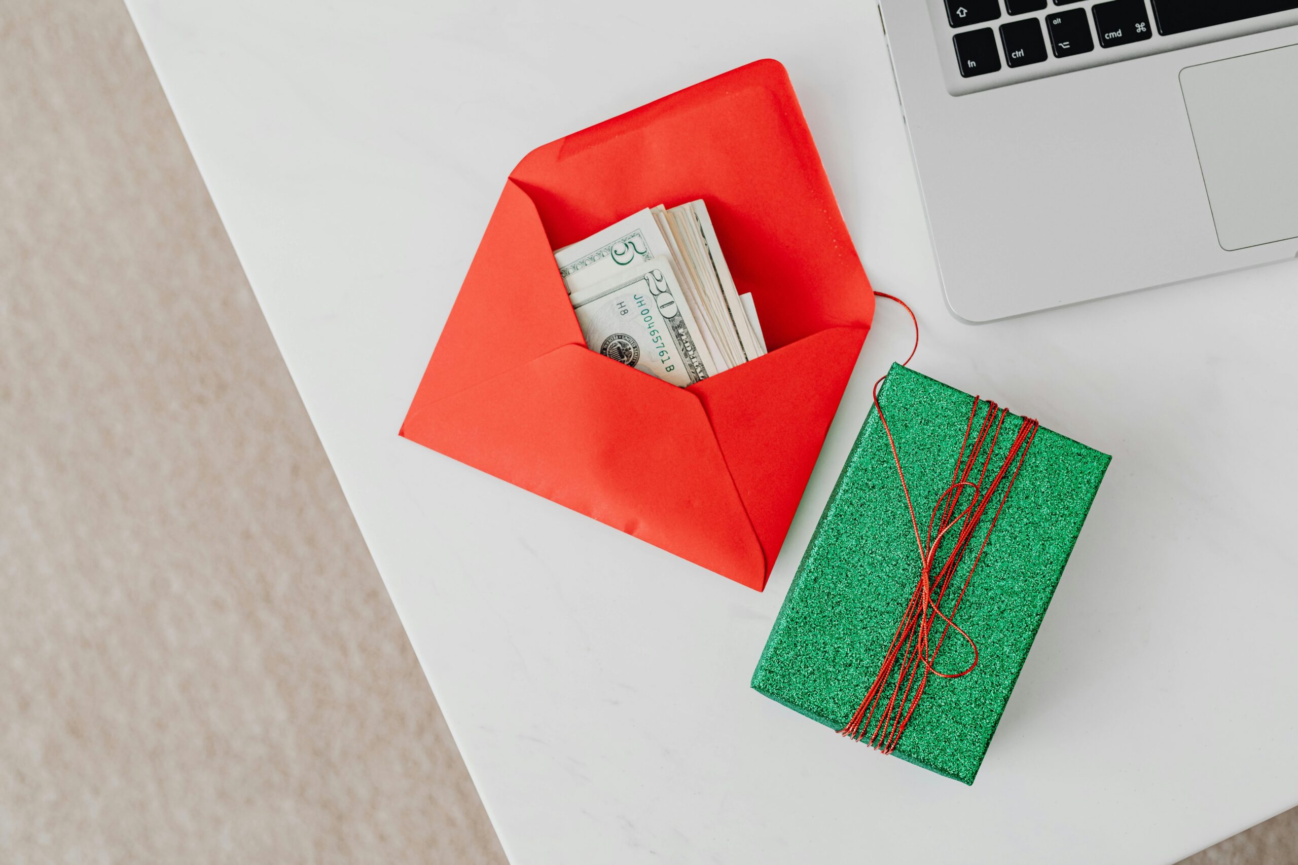 A green gift box and red envelope with cash on a desk beside a laptop, emphasizing holiday or financial themes.