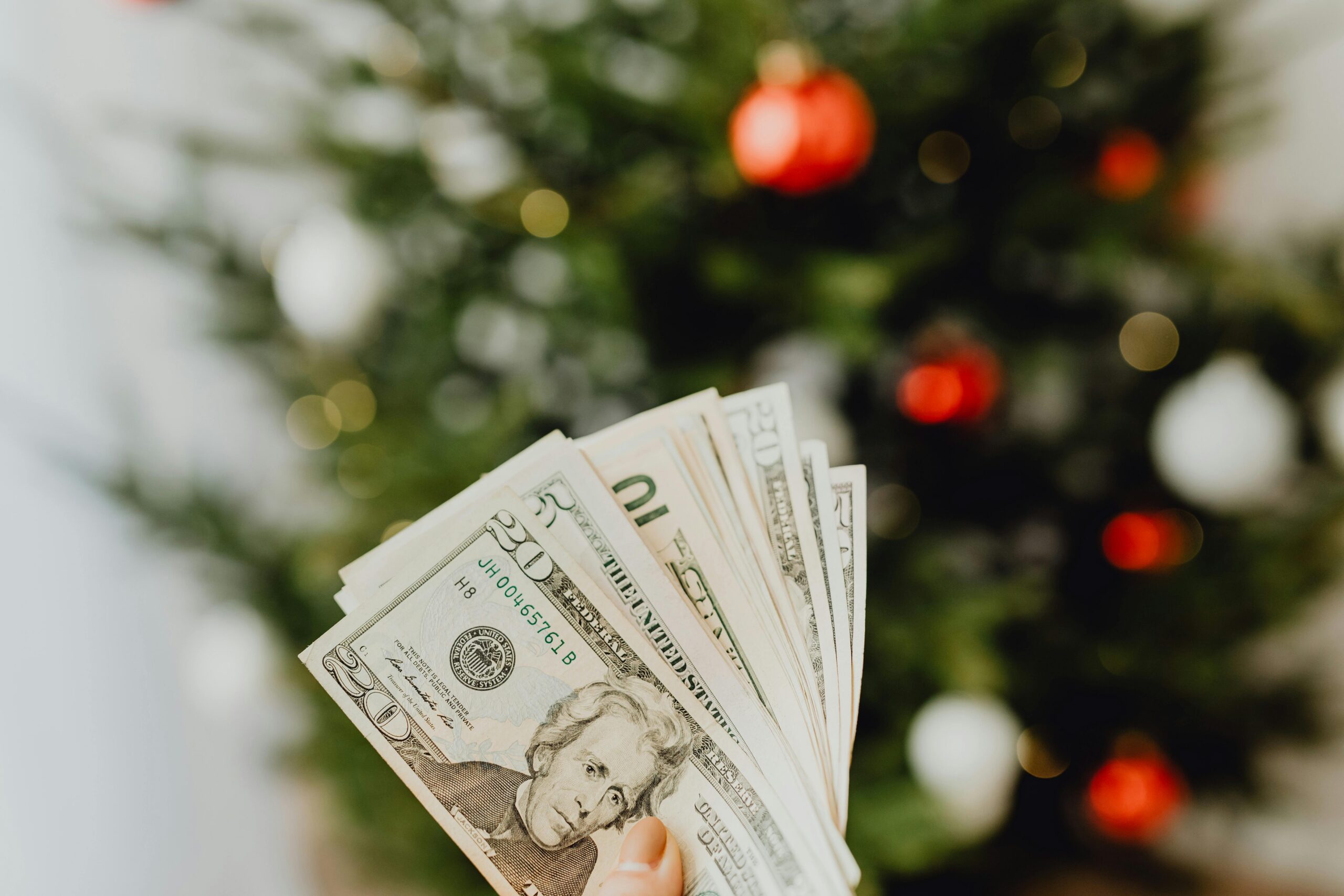 A close-up of a hand holding dollar bills with a blurred Christmas tree in the background.