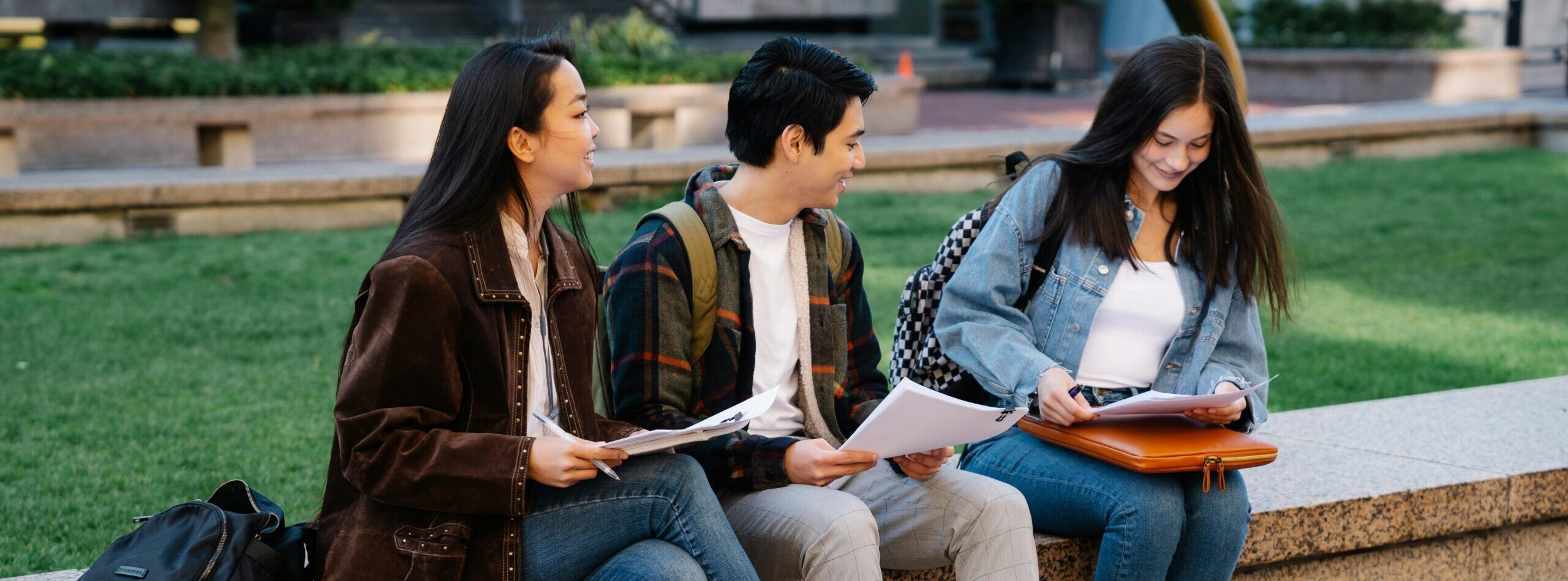 Three college students engaged in study session on a campus bench, enjoying a sunny day.